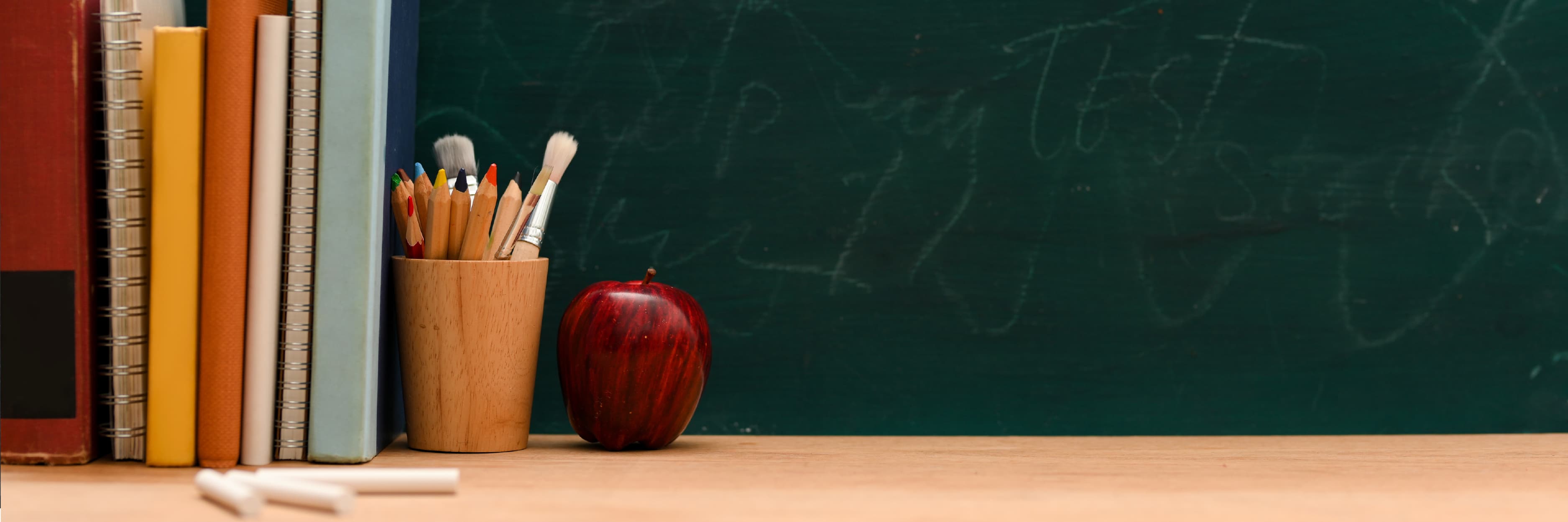A desk with books, chalk, crayons, and an apple on top, and a green chalkboard behind it.