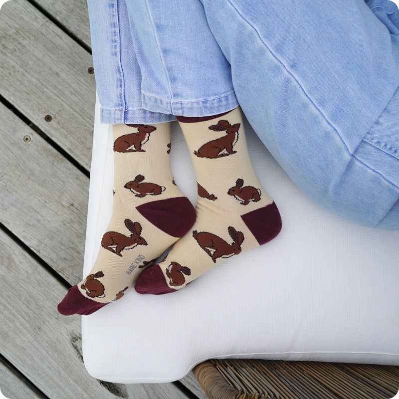 A person wearing beige socks with a brown hare pattern and dark burgundy toe/heel accents, paired with light blue jeans, sitting on a white cushion over a wooden deck.