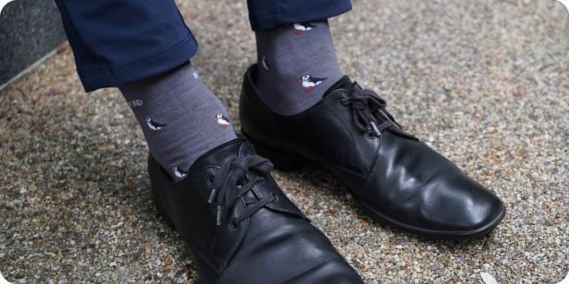 Gray crew socks with a small duck print, worn with black leather dress shoes and navy blue trousers, standing on a textured concrete surface.