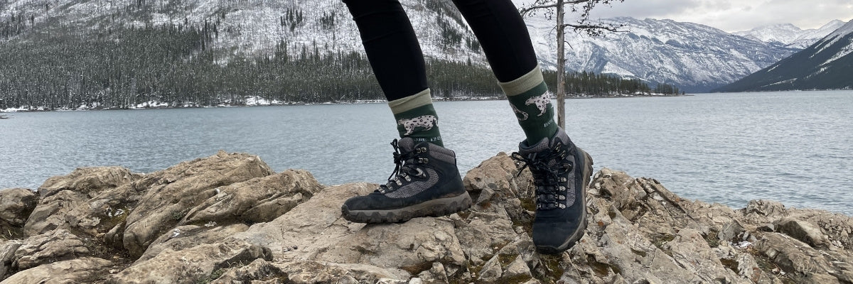 Person wearing hiking boots and socks on a rocky outcrop with a scenic lake and mountains in the background