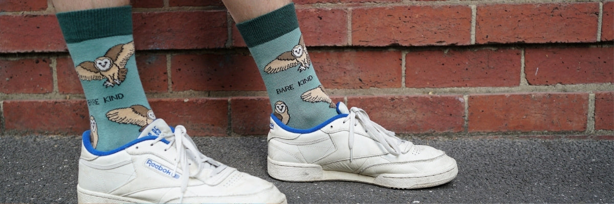 A person wearing light blue crew socks with owl illustrations and white sneakers, standing against a red brick wall.