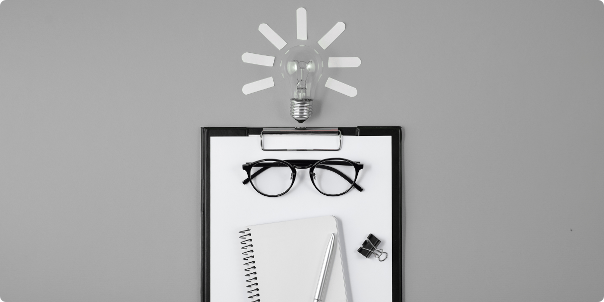 Clipboard with glasses, pen, and notebook on a gray background with a light bulb icon above.
