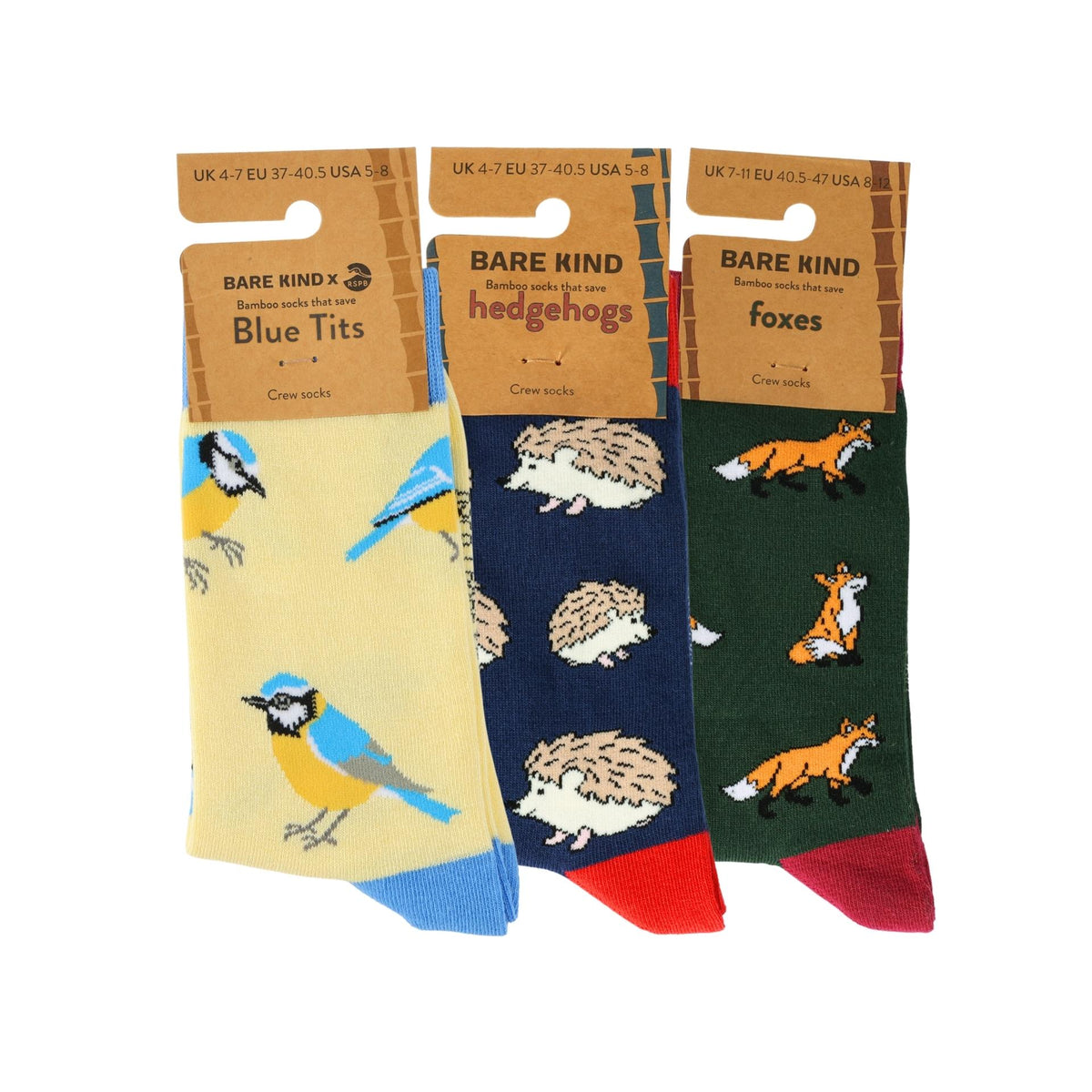 Three pairs of Bare Kind socks displayed side-by-side on their cardboard hangers against a white background. The left pair is 'Blue Tits' in pale yellow with blue and yellow birds; the middle pair is 'hedgehogs' in navy blue with brown hedgehogs and a red toe; the right pair is 'foxes' in dark green with orange and white foxes and a red toe.