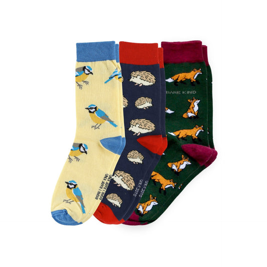 Three pairs of patterned crew socks displayed together on a white background. The left sock is pale yellow with blue birds; the middle sock is navy with hedgehogs and red cuffs/soles; the right sock is dark green with orange foxes and burgundy cuffs/soles.
