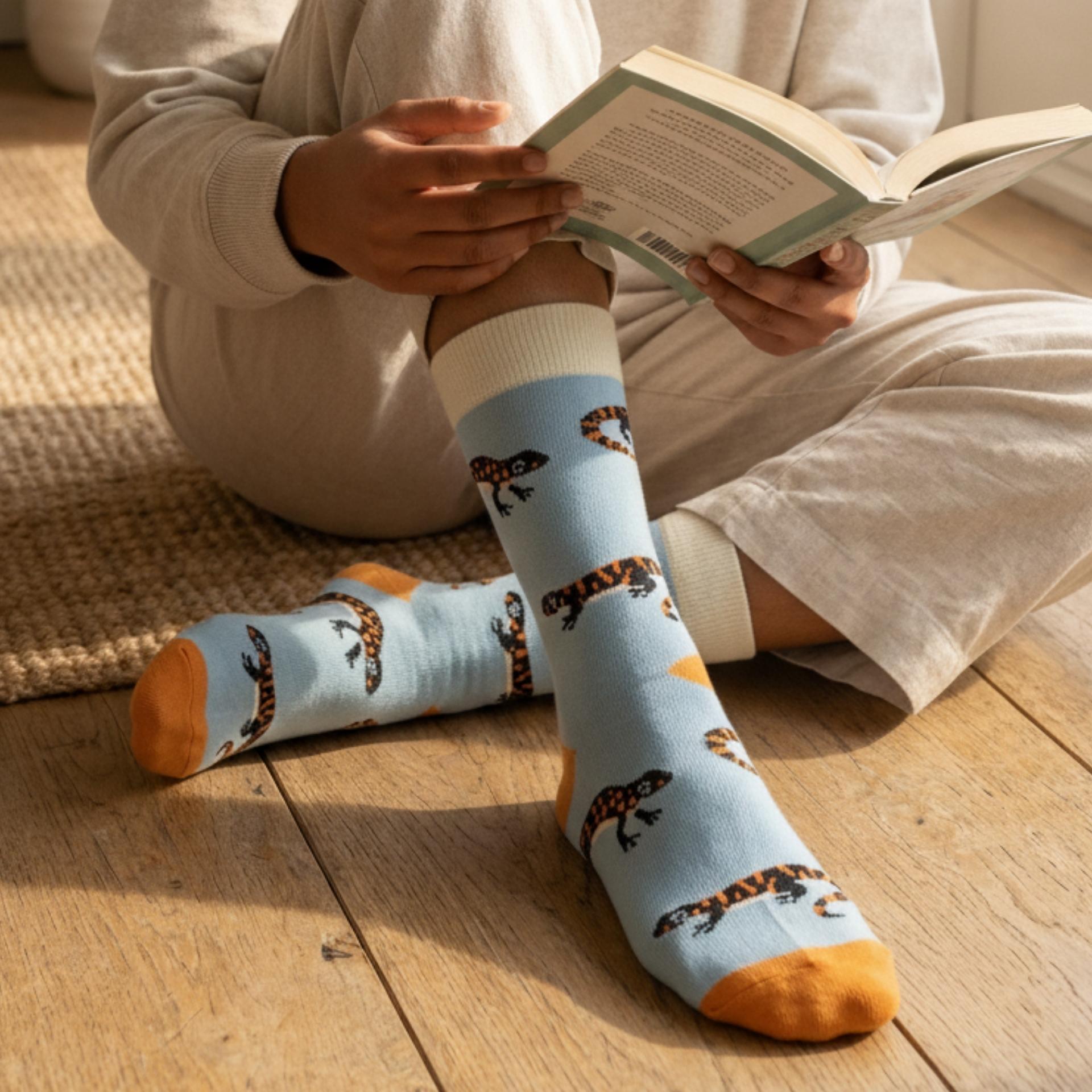 Person wearing blue socks with salamander pattern sitting on a wooden floor reading a book.