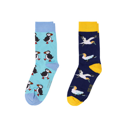 Two novelty crew socks shown flat against a white background. The left sock is light blue with puffin illustrations, and the right sock is dark navy with gannet illustrations and yellow accents.