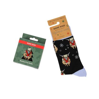 Bare Kind bamboo crew socks shown with their green packaging box on a white background. The socks are black with a pattern of Highland cows wearing red scarves and white snowflakes, with a light blue toe.