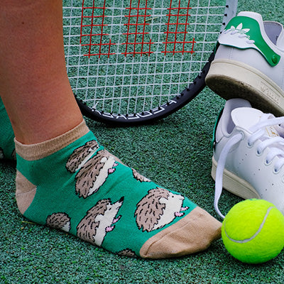 Person wearing green trainer socks with hedgehog pattern on a tennis court, next to a tennis racket and ball.