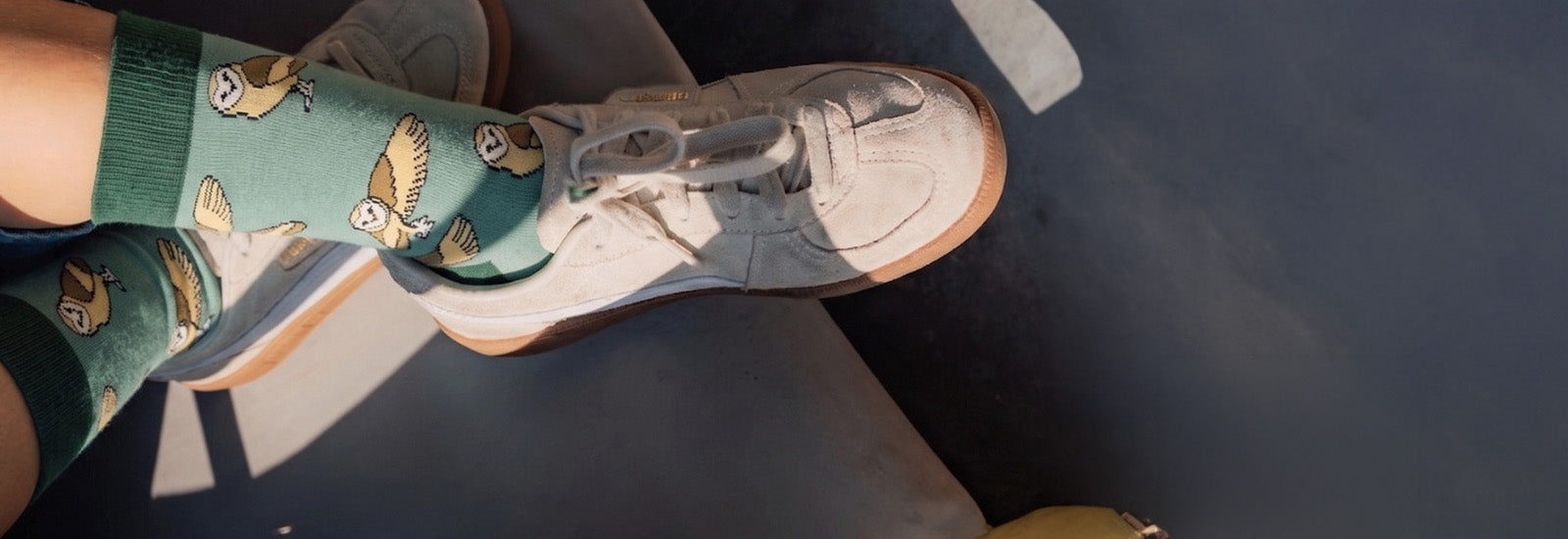 A close-up lifestyle shot of a person's feet wearing green socks with a barn owl pattern and off-white sneakers, resting on a dark surface.
