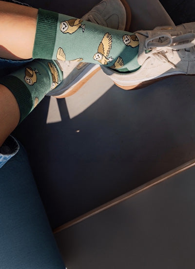 A person wearing green crew socks with a barn owl pattern, paired with white sneakers, seated on a dark train or bus.