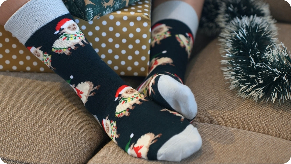 A person wearing black christmas sheep socks, sitting on a brown fabric surface with a gold polka-dot gift box and tinsel in the background.