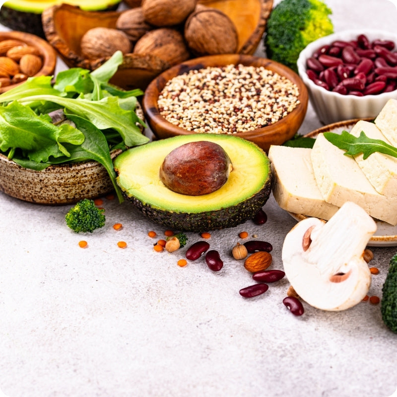 Assorted healthy foods including avocado, nuts, seeds, and vegetables on a light surface.