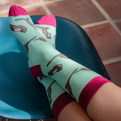 A person's feet wearing mint green socks with a brown otter pattern, bright pink toes and heels, and ribbed pink cuffs, resting on a teal chair over a tiled floor.