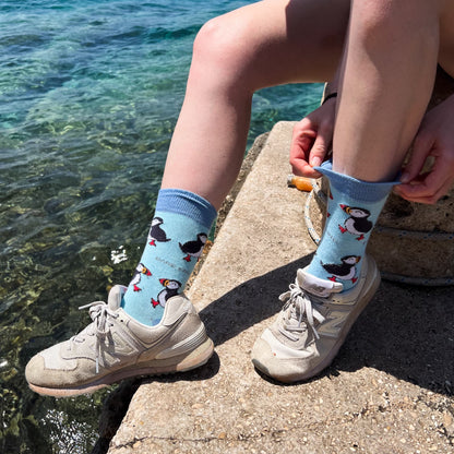 lifestyle image of model putting on blue puffin bamboo socks next to the ocean