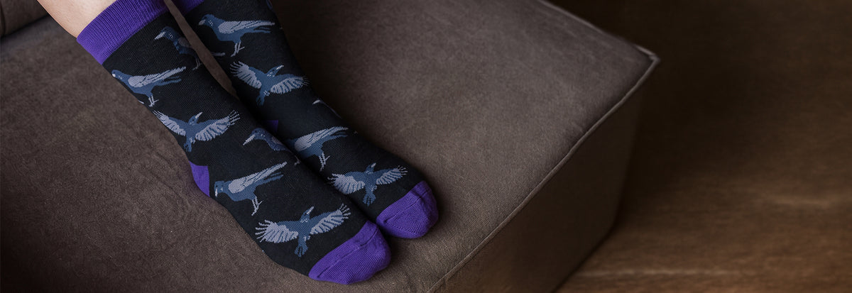 A pair of crew socks with a dark charcoal background, featuring a pattern of ravens and solid purple toes and cuffs, shown worn on feet resting on a brown fabric surface.