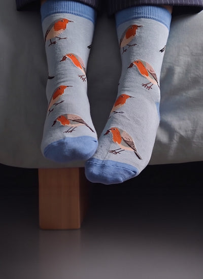 A pair of light blue crew socks with a pattern of orange birds, shown on a person's feet resting on a bed.