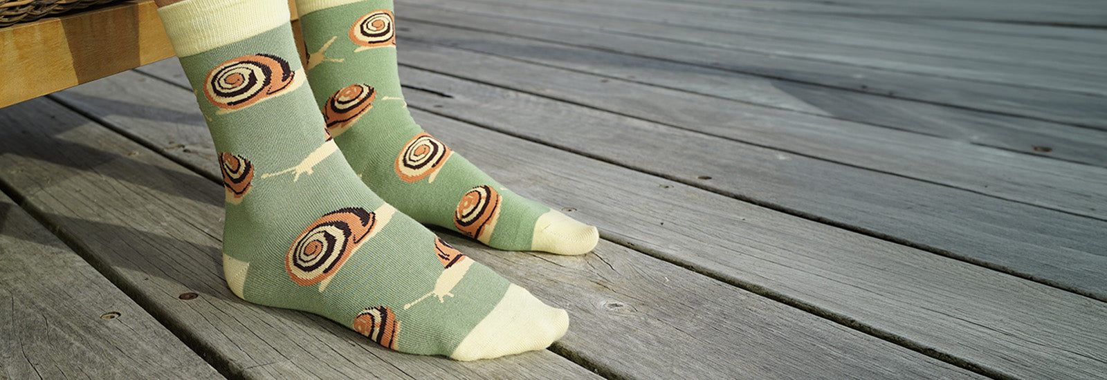 A pair of light sage green crew socks with a brown and black spiral shell pattern and cream tips, worn on feet standing on weathered gray wooden deck planks.