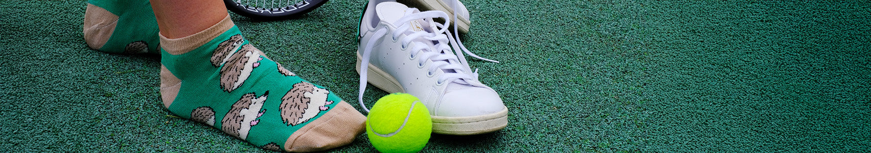 Person wearing green trainer socks with hedgehog pattern on a tennis court, next to a tennis racket and ball.