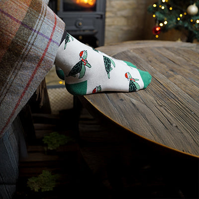 Person wearing a festive turtle socks with holly design on a wooden surface, Christmas decorations in the background.