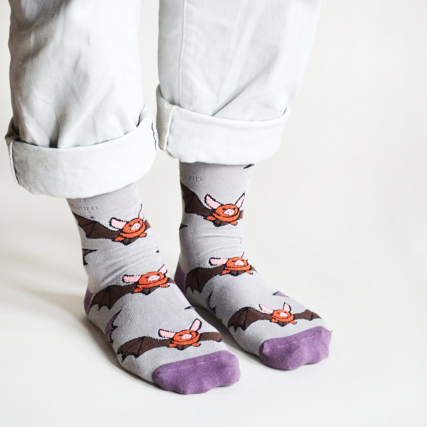 Side angle view of a model with both feet flat on the floor as the model stands, wearing purple and grey bat bamboo socks